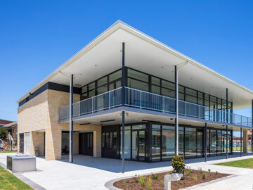 A modern two-story building with large glass windows, a wide balcony, and a flat roof under a clear blue sky. The exterior features stone and metal elements, with landscaped greenery in the foreground.