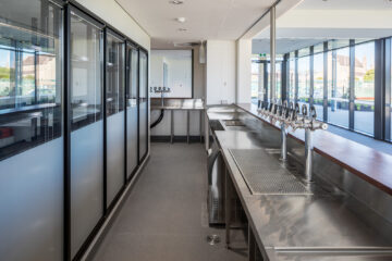 A modern commercial bar setup with stainless steel countertops, multiple beer taps, glass-door refrigerators, and large windows providing a view outside. The area is clean, bright, and empty of people.