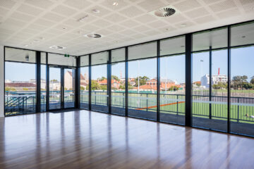 A spacious room with floor-to-ceiling glass windows overlooking a large outdoor sports field and buildings in the distance, with polished wooden floors and a modern ceiling.