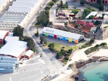 Aerial view of a coastal area with a large building featuring colorful panels, surrounded by roads, smaller buildings, trees, and a sandy beach next to clear water.