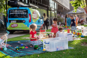 Children play with toys on grass next to large plastic bins and a blue van marked “Buster.” Adults and buildings are in the background, and the scene is bright and sunny, suggesting a community outdoor event.
