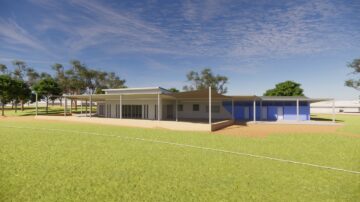 Modern one-story building with large windows, flat roofs, and shaded walkways sits on a grassy field, surrounded by trees under a blue sky with wispy clouds.