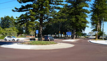 A small roundabout with cars driving around it, surrounded by tall pine trees and street signs, on a sunny day. The sky is clear and blue, and there are benches and a park area in the background.