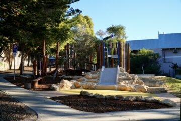 A playground with a climbing structure, slide, rocks, wooden poles, and trees surrounding it. There is a sidewalk, bench, and buildings in the background. The area is well-lit by sunlight.