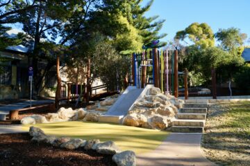 A colorful playground with a slide, rock steps, and climbing structures surrounded by trees and greenery on a sunny day.