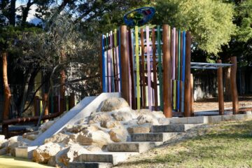 A colorful playground structure with wooden poles, a small rock climbing area, and steps, surrounded by trees and sunlight filtering through the leaves.