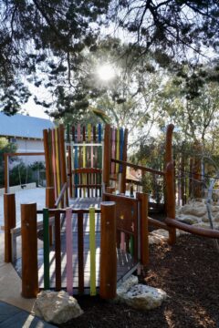 A colorful wooden playground structure is shaded by trees, with sunlight filtering through the branches, surrounded by rocks and mulch.