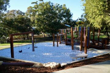 A playground with swings, a climbing structure, and a slide, all set in a sandy area surrounded by trees and grassy parkland on a sunny day.