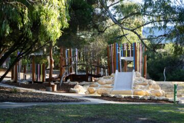 A colorful playground with wooden structures, a slide, and climbing rocks is surrounded by trees and shaded areas. Sunlight filters through the branches onto the play area and curved walking paths.