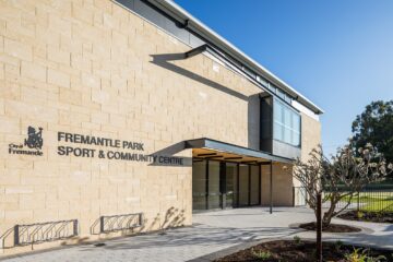 Modern building with light-colored stone walls and large windows. Sign reads Fremantle Park Sport & Community Centre. Small trees and bike racks are outside, with clear blue sky in the background.