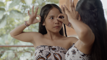 Two young women in patterned clothing face each other, performing a traditional dance gesture with their hands creating circles near their eyes. The background is blurred with greenery and soft light.