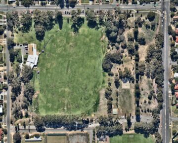 Aerial view of a large sports complex with multiple grassy fields, buildings, parking lots, and surrounding trees, bordered by roads and residential areas.