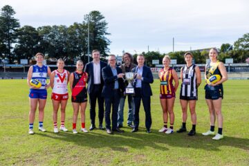 Eight women in football uniforms and four men in suits stand on a grassy sports field, smiling and holding a trophy in the center of the group. The women each wear different team colors.