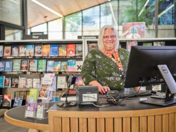 A smiling librarian stands behind a circular wooden desk with a computer in a modern library, surrounded by bookshelves filled with colorful books.