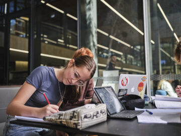 A young woman with glasses writes in a notebook at a table with a tablet, laptop, and books. She is studying outdoors near glass windows, with another person and papers visible nearby.