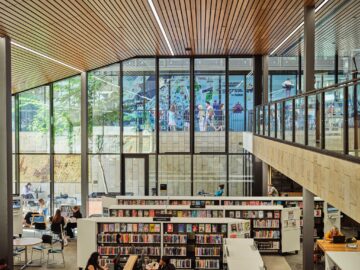 Modern library interior with tall windows, wooden ceiling, bookshelves, and people reading or working at tables. A colorful mural and people walking are visible outside through the large glass windows.