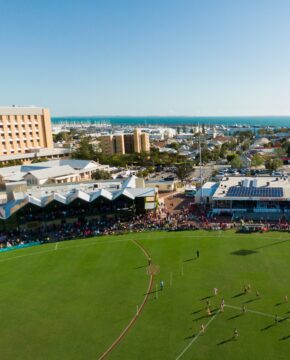 Aerial view of an outdoor sports stadium with a game in progress, spectators in the stands, and a coastal town with buildings and a marina in the background under a clear blue sky.