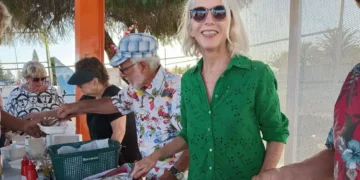 A smiling woman in a green shirt stands in the foreground, holding tongs. Behind her, people serve food at an outdoor event on a sunny day. Others wear casual, colorful clothes and hats, and theres a tree providing shade.