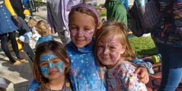 Three young children with colorful face paint smile and pose together outdoors at a sunny event, surrounded by other people and children in the background.