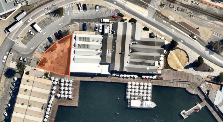 Aerial view of a waterfront building complex with a dock and outdoor seating areas. A triangular section of land is highlighted in orange near a curved road labeled Mews Rd.