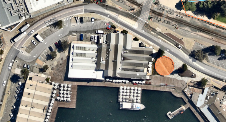 Aerial view of a waterfront area with buildings, parked cars, a docked boat, and a circular orange section on the ground beside a road, near the edge of the water.