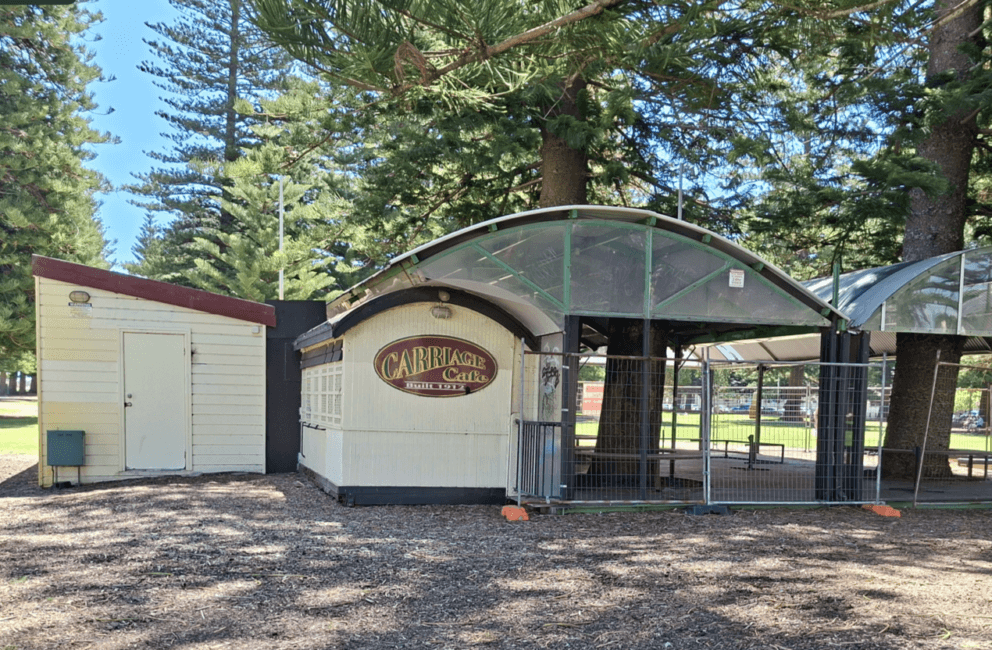A closed café kiosk with a curved roof and a Carriage Café sign, next to a small white shed, surrounded by tall pine trees and fencing in a park setting.