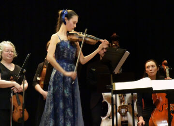 A woman in a blue dress plays violin on stage with other musicians holding string instruments, while sheet music rests on stands nearby.