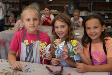 Three young girls sit at a table in an art classroom, smiling and holding colorful mosaic crafts shaped like people. Other children and art supplies are visible in the background.