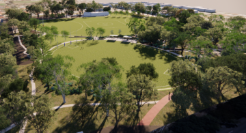 Aerial view of a circular sports field surrounded by trees, walking paths, and some people. There are white markings for sports and buildings visible in the background.