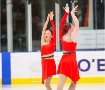 Two figure skaters in matching red dresses and white skates perform a routine on the ice, both raising their arms gracefully, with one facing forward and the other facing backward.
