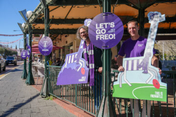 Two smiling men in purple shirts hold Fremantle Dockers signs outside a café with outdoor seating, supporting their team. The signs read LETS GO FREO! and THIS IS FREMANTLE..