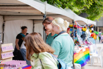 Three people look at jewelry at an outdoor market stall, with a rainbow flag visible on a nearby table and other people browsing in the background.