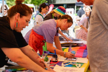 People gather around a colorful table outdoors, making art with fabric, markers, and paints. One person in a rainbow hat looks on as another paints, while others chat and participate in the creative activity.