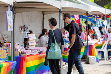 Two people browse jewelry at a market stall decorated with rainbow Pride flags and colorful tablecloths. Other vendors and rainbow decorations are visible in the background.