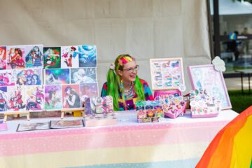 A smiling person with colorful hair and accessories sits behind a booth displaying vibrant art prints, buttons, and stickers at an outdoor event. The table is covered with a pastel cloth and bright, whimsical artwork.