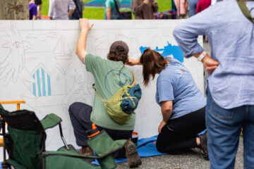 Two people kneel on the ground, painting a large mural with outlined shapes of houses and birds. Supplies and a green chair are nearby. Other people stand around, watching the artwork progress.