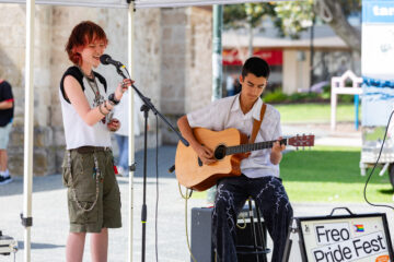 Two young musicians perform at an outdoor event—one singing into a microphone, the other playing an acoustic guitar—next to a sign reading Freo Pride Fest with rainbow and trans flags.