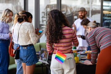 A group of people gather around a table with art supplies. One person has a rainbow pride flag in their pocket. Some people are painting or writing, and the scene appears to be outdoors near a building.