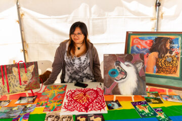 A woman stands behind a colorful table displaying her artwork, including paintings of animals and a woman, at an outdoor art fair. A white tent serves as the background.