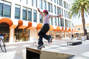 A person wearing a helmet and roller skates performs a trick on a rail in an outdoor urban area with modern buildings and palm trees.