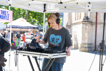 A DJ wearing headphones and a grey t-shirt is mixing music outdoors under a canopy at a daytime event, with people and a stone building visible in the background.