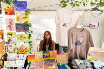 A person with curly hair and glasses sits at a colorful booth selling art prints and illustrated t-shirts at an outdoor market, surrounded by vibrant artwork and greenery.