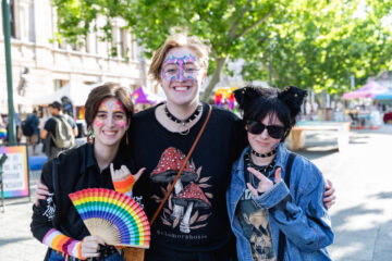 Three smiling people outdoors at a festival, wearing colorful face paint and alternative fashion. One holds a rainbow fan, and trees with green leaves are in the background.