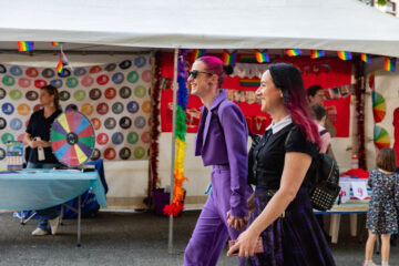 Two people holding hands and smiling walk past a colorful booth decorated with rainbow flags and a game wheel at an outdoor event. Other people and a child are visible near the booth.