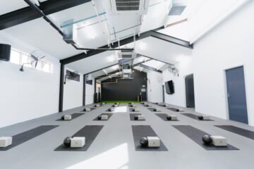 A bright, modern fitness studio with rows of black yoga mats, each set up with a block and a kettlebell, ready for a group exercise or yoga class. The room has high ceilings and white walls.