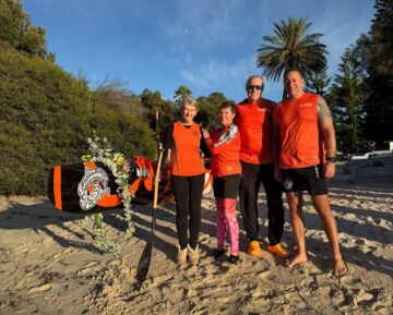 Four people in orange sportswear stand on a sandy beach beside a decorated paddleboard, with trees and a blue sky in the background. One person holds a paddle; all are smiling at the camera.