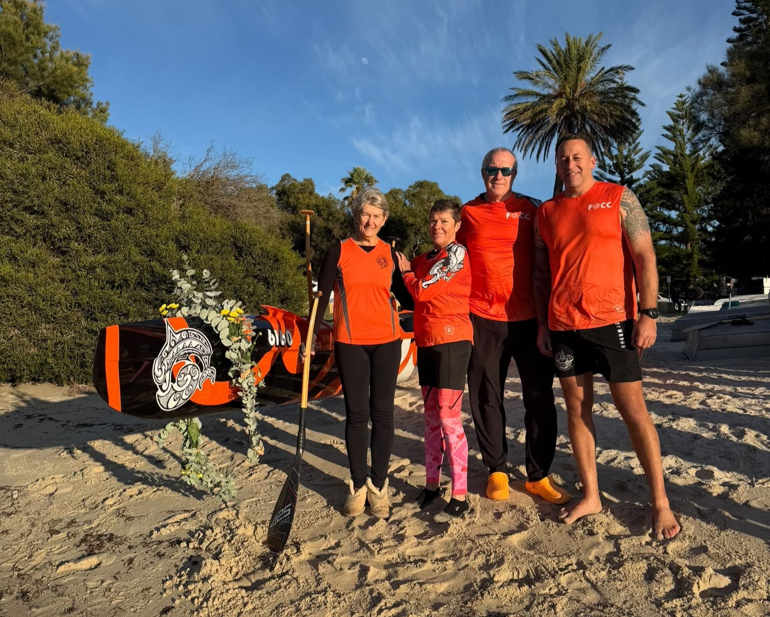 Four people in orange sportswear stand on a sandy beach beside a decorated paddleboard, with trees and a blue sky in the background. One person holds a paddle; all are smiling at the camera.
