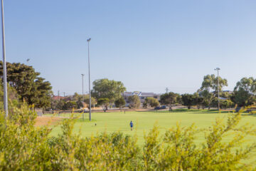 A person walks across a large, green sports field surrounded by trees and light poles, with houses and clear blue sky in the background.
