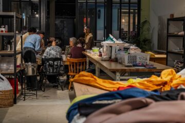 A group of people sit around a table working together in a craft studio filled with supplies, baskets, shelves, and fabrics in the foreground.