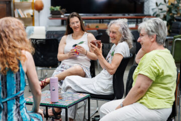 Four women sit in a circle indoors, chatting and laughing. One holds a mug, another a small object. They appear relaxed and engaged, seated around a colorful table in a bright, welcoming room.